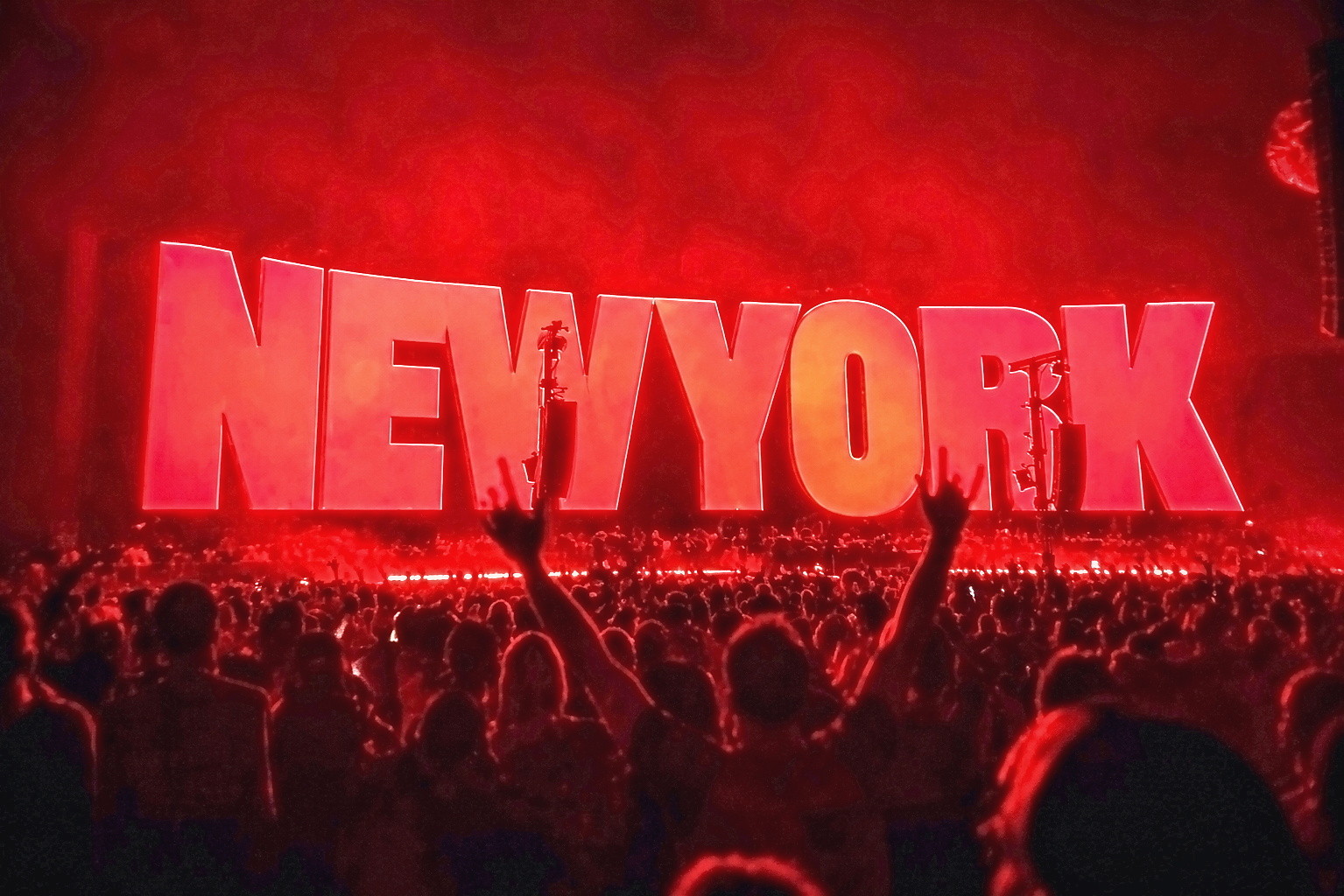 A large crowd with raised hands beneath a giant illuminated NEW YORK sign